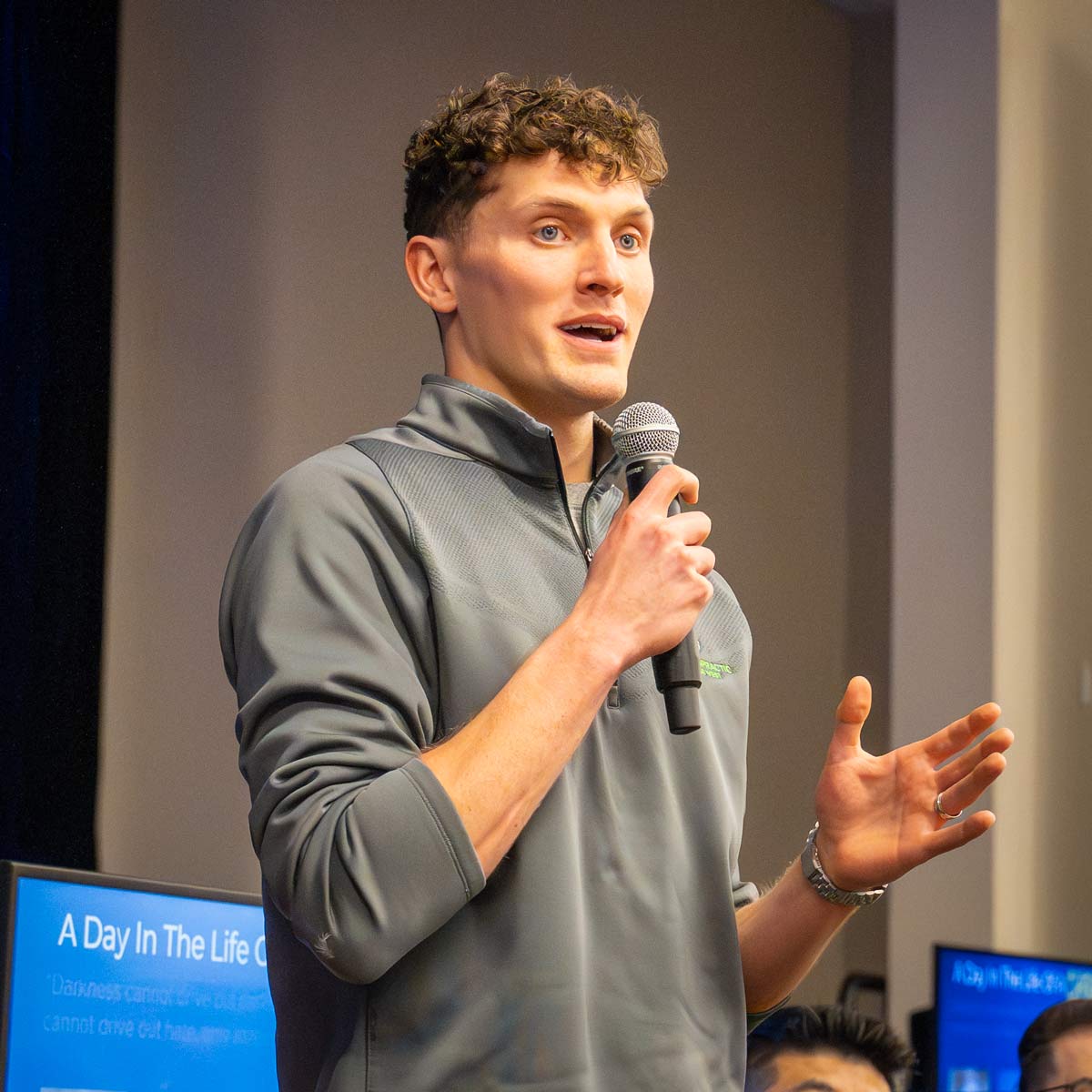 Young man speaking into a handheld microphone on stage, wearing a gray athletic pullover and gesturing with his other hand.